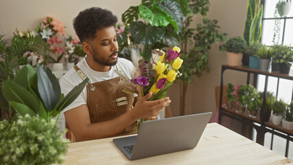 A handsome man arranges flowers in a green indoor florist shop, with a laptop on his wooden table.