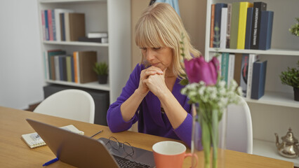 A thoughtful woman working from home, with a laptop, glasses, and flowers on her table.