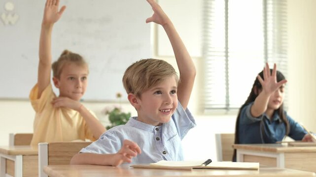 Caucasian boy raising hand for asking and answering teacher surrounded by diverse children studying at classroom. Happy student working together, voting, volunteering, calling instructor. Pedagogy.