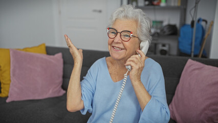 A cheerful senior woman in glasses speaking on a landline phone in a cozy living room, expressing happiness and conversation.