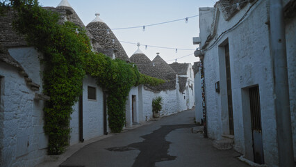 A charming narrow street in alberobello, italy, showcasing traditional white trulli houses with grey conical roofs and lush green ivy climbing the walls under twilight skies.