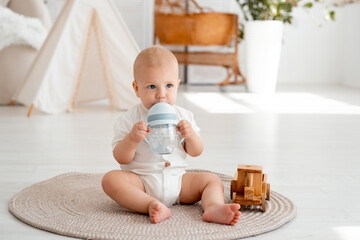 a baby drinks water from a bottle or a drinking cup sitting on the floor in a bright bedroom, a...