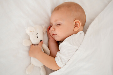 a newborn baby sleeps sweetly holding a toy in his hands, a place for text, a baby sleeps on a white bed under a blanket with a pen under his cheek, a healthy baby's sleep