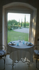 A serene setup of a romantic table for two with neatly set plates and glasses in an italian restaurant, offering a picturesque view of lush greenery through a large window.