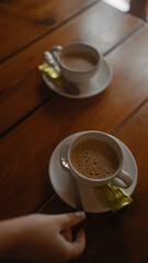 Person reaches for a chocolate with coffee in focus on a wooden table, invoking a cozy cafeteria setting.