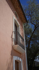 Spanish villa balcony with intricate railing under a vibrant blue sky, framed by a lush green tree and casting soft shadows.