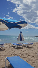 Sunny beach scene with blue loungers and umbrellas, clear sky, and sparkling sea, implying leisure and vacation.