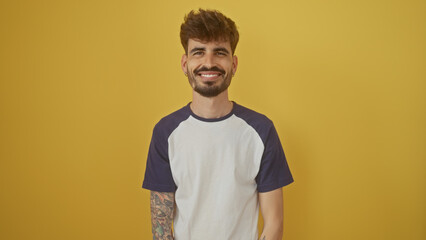 Handsome young man with beard and tattoos smiling confidently against a vibrant yellow background