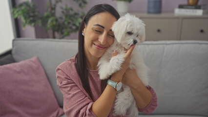 A smiling young woman affectionately holding a fluffy white bichon maltese indoors in a cozy living room.