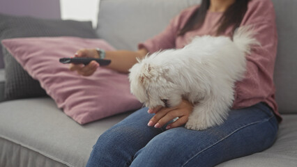 A young woman relaxes on a sofa with a fluffy bichon maltes on her lap while holding a remote in a...