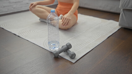 A woman in sportswear sits on a yoga mat near a water bottle and dumbbells in a home interior.