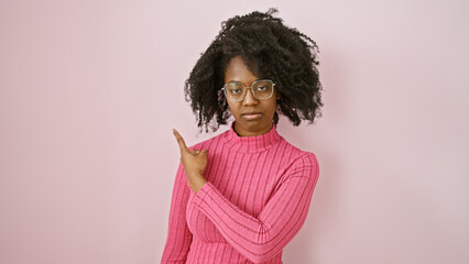 African american woman in glasses pointing sideways in a pink sweater against a plain background.