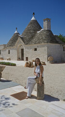 A young hispanic woman sitting outside a traditional trulli house in the old town of alberobello,...