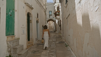 Young hispanic woman in a white dress walking through the old town streets of ostuni, puglia, italy, with a rustic wall and green door in the background.