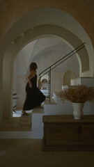 A young hispanic woman in a black dress ascends a staircase inside a traditional italian masseria,...