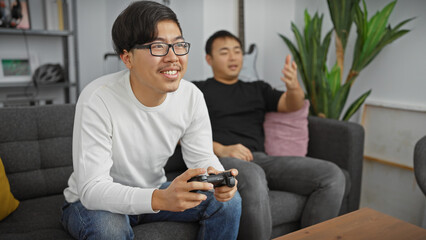 Obraz premium Two asian men enjoying time together playing video games in a cozy living room setting.