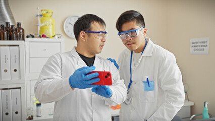 Two men scientists in lab coats and safety goggles analyzing data on a smartphone in a laboratory setting.