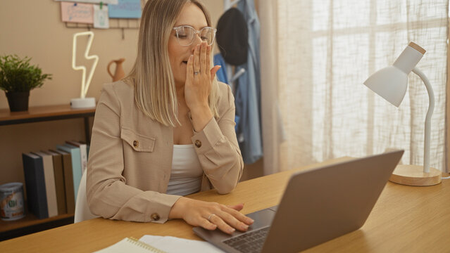 A beautiful adult blonde woman in a home office yawning while working on a laptop in a bright indoor living room setting.