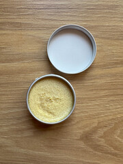 Small container of yellow scrub is placed on a wooden surface with its lid open. Texture scrub for face. Cosmetic product and skincare concept