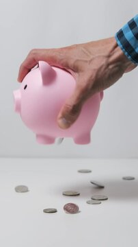 Vertical video. Man empties piggy bank, coins scatter across the table, symbolizing financial management