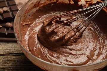 Chocolate dough in bowl and whisk on wooden table, closeup