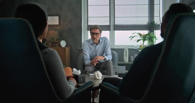 Mature psychotherapist talks to young family, provides couples therapy. Back view of multiethnic couple sitting on chairs, listening psychologist on counseling session. Psychology and sexology concept