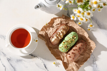 Delicious eclairs, cup of tea and chamomile flowers on light background