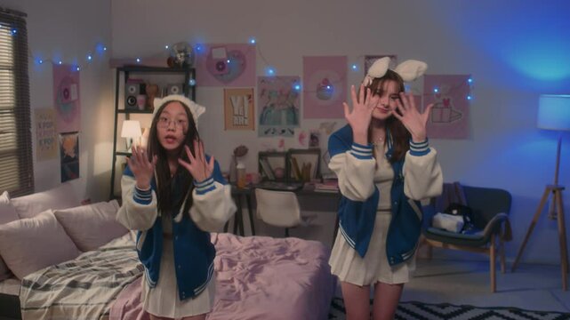 Two teenage girls in matching outfits dancing to k-pop song and looking at camera in bedroom