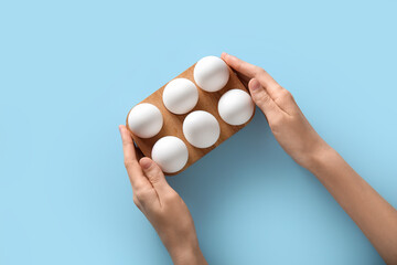 Female hands with wooden holder of raw chicken eggs on blue background