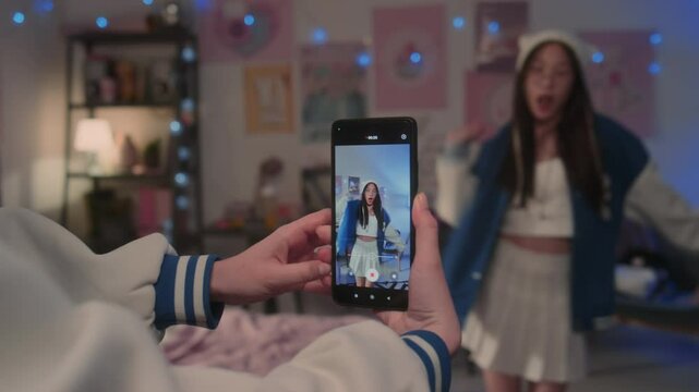 Close-up of hands of unknown teenager holding smartphone and recording vertical video of Korean girl dancing to k-pop song in bedroom