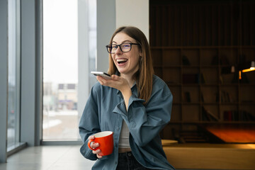 Cheerful young woman in glasses recording voice message in library.
