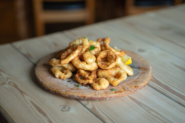 Calamari in a crispy golden batter served on a circular wooden bread board on a rustic wooden table with a piece of lemon. The focus is shallow.