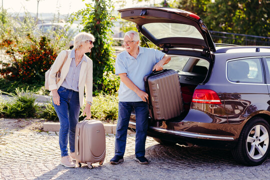 An elderly couple loads luggage into the trunk of their car. The man lifts a suitcase while the woman stands by, holding another suitcase, preparing for or after a trip.