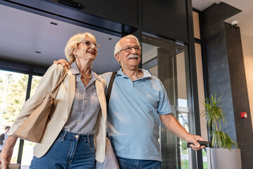An elderly couple enters a hotel lobby, pulling their suitcases. They are dressed casually,...