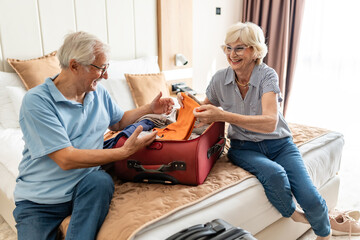 An elderly couple sitting on a hotel bed, smiling as they unpack their suitcase together,...