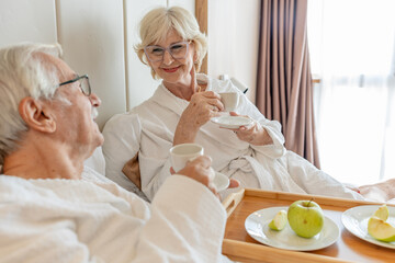 An elderly couple enjoys breakfast in bed, wearing white bathrobes, and sipping coffee. A tray with...