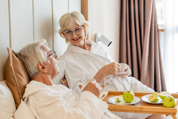 An elderly couple enjoys breakfast in bed, wearing white bathrobes, and sipping coffee. A tray with...