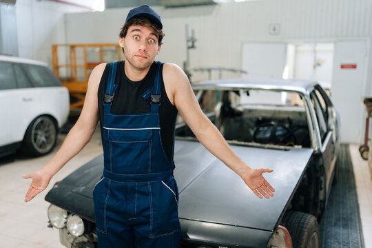 Shocked doubtful repairman in workwear clothes, trying to solve problem. Puzzled mechanic male in uniform shrugging shoulders while not knowing what disrepair with repair car. Concept of car service.