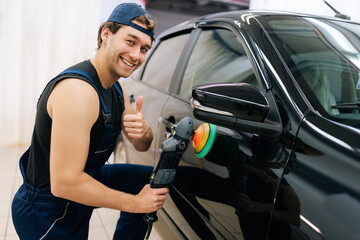 Friendly mechanic male in uniform polish auto door using grinding unit in automobile repair and renew service station, smiling looking at camera. Concept of car repair and maintenance work