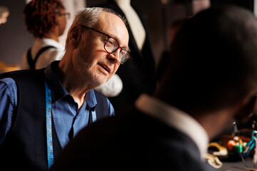 Close up of master suitmaker checking african american apprentice sartorial garment sketches in...