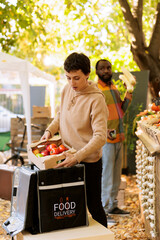 Female employee with backpack delivering seasonal bio products in box, preparing to take natural food order to customers. Woman delivery courier picking up produce from local market stall.