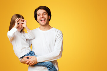 Naklejka premium Funny little daughter and daddy fooling, having fun over yellow background, girl imitating binoculars