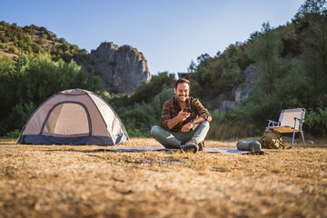 Fototapeta premium Adult man sit near tent and use mobile phone on camping trip