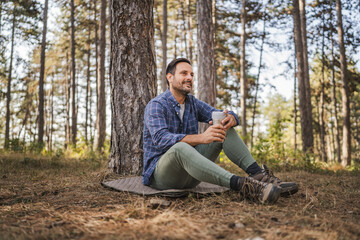 Adult man sit on mat in front tree and drink coffee from thermos