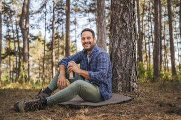 Obraz premium Adult man sit on mat in front tree and drink coffee from thermos