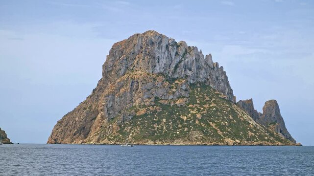 Daytime close up footage of Es Vedra island, off the coast of Ibiza, taken from a sailing boat, with blue skies and and a few clouds visible on the horizon