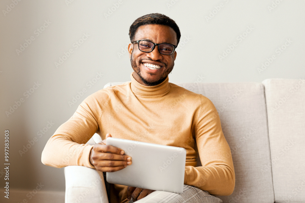 Poster young afro man holding digital tablet computer and smiling at camera sitting on couch indoor. select