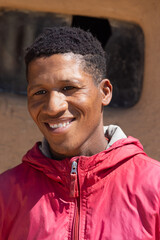 african village, young man with toothy smile, half portrait head and shoulders , standing in front of a mud house wall