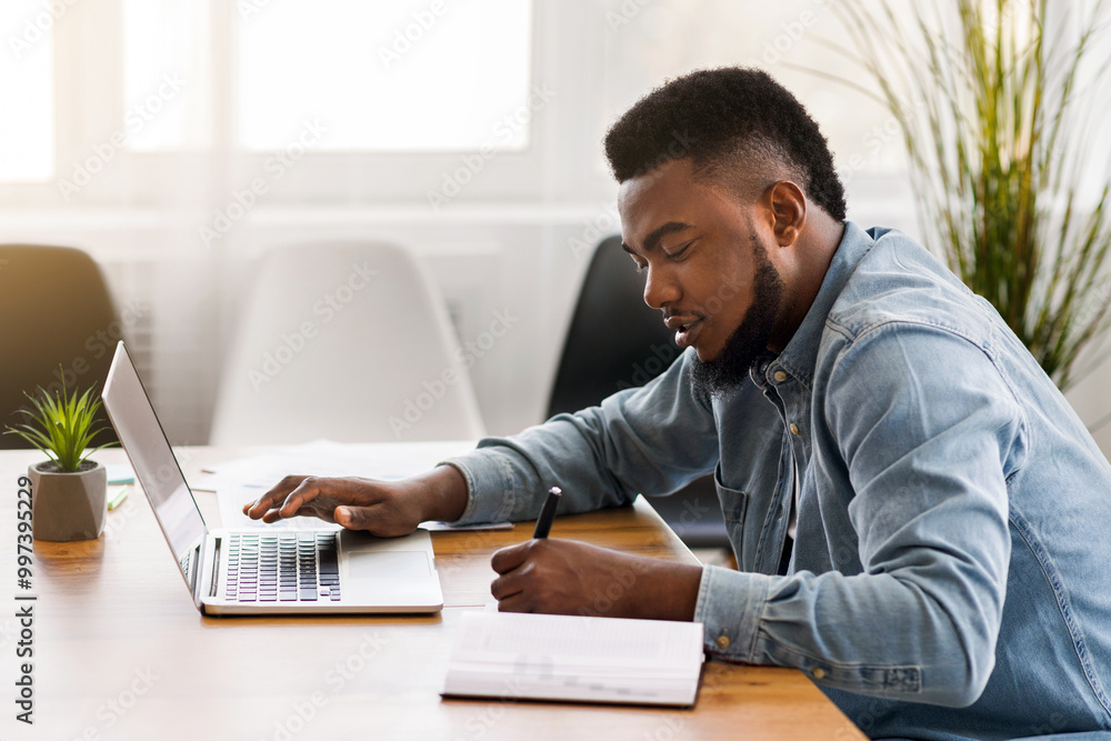 Poster african american employee using laptop and taking notes while working in office, side view with copy