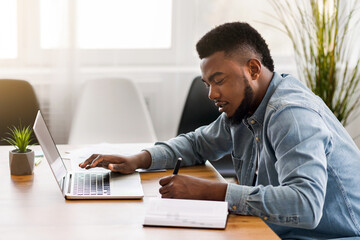 African American employee using laptop and taking notes while working in office, side view with copy space